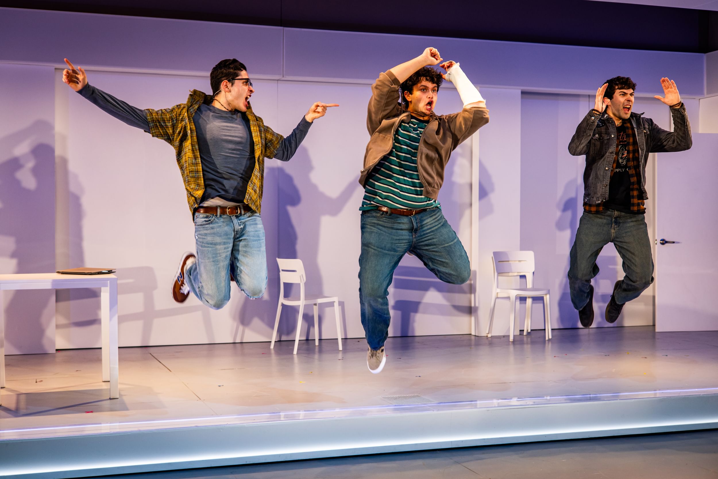 The three young male actors, in casual contemporary clothes, are caught jumping into the air in dramatic poses.