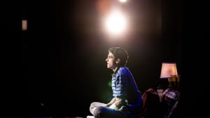 Norman, a young white man in striped shirt & cast on one arm, sits in profile on a bed, under a bright spotlight in the dark.