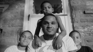 Three young Black boys sit around a young Black man in front of a white door on a porch, looking at camera. Black and white