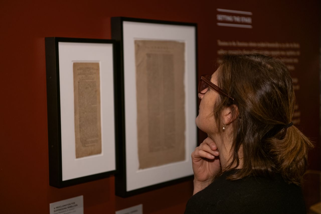 Close-up on a white woman with brown hair, seen from behind as she looks closely at framed 18th-century broadsides.