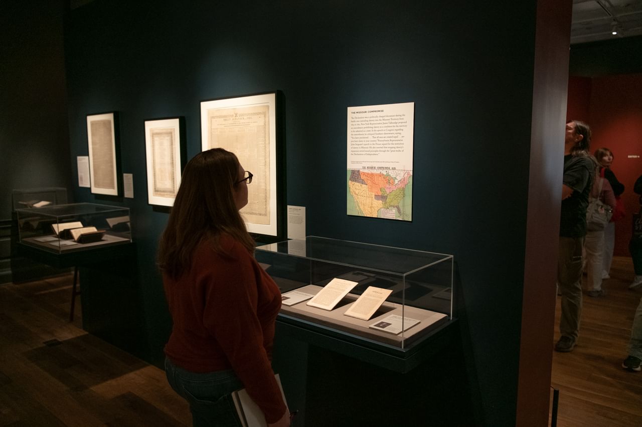 Alaina, a white woman with blond hair, wearing an orange sweater, looks at a historical map in the gallery.