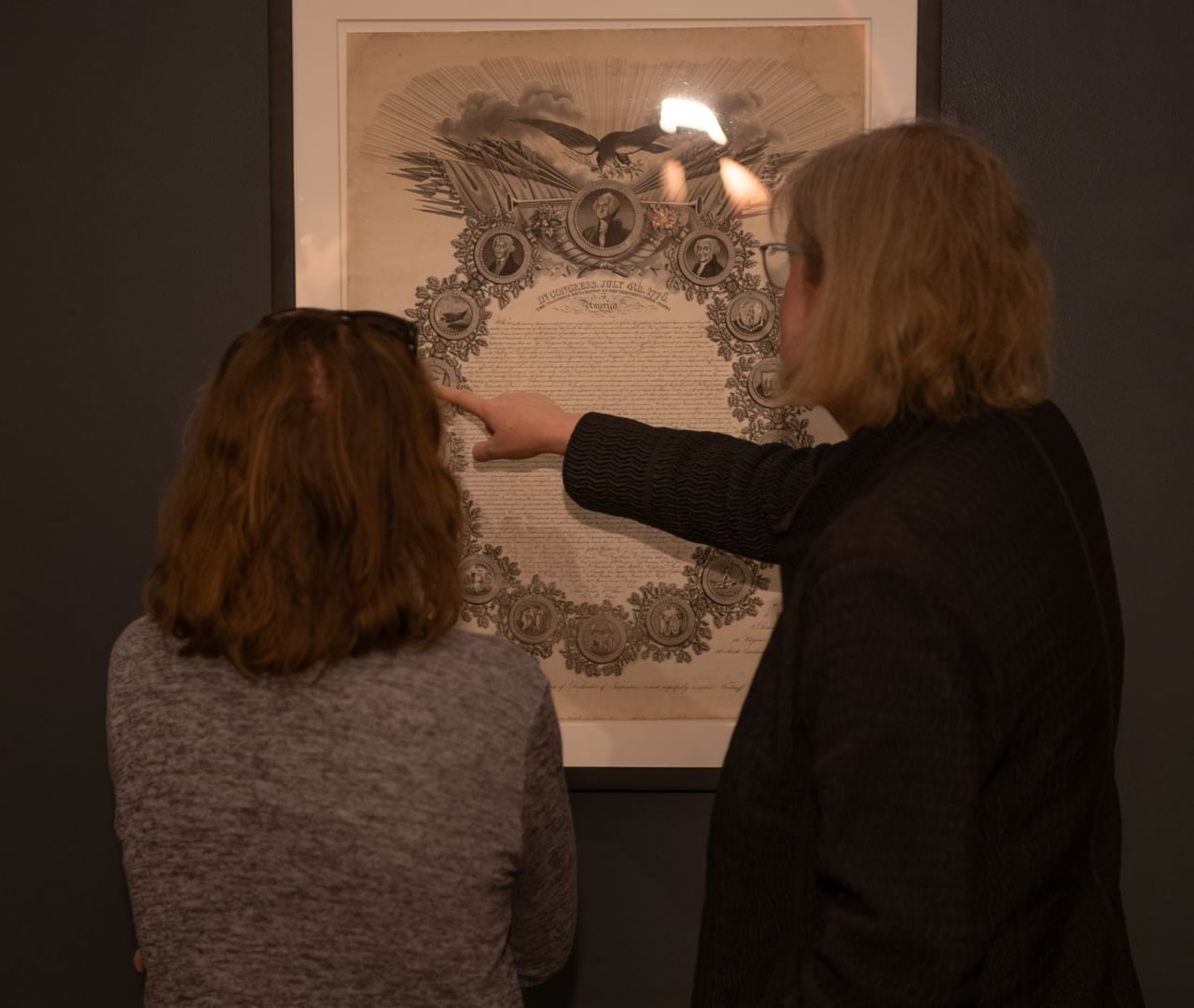 Seen from behind, two women, one of them pointing to the document, look closely at a poster-size 1819 printing.