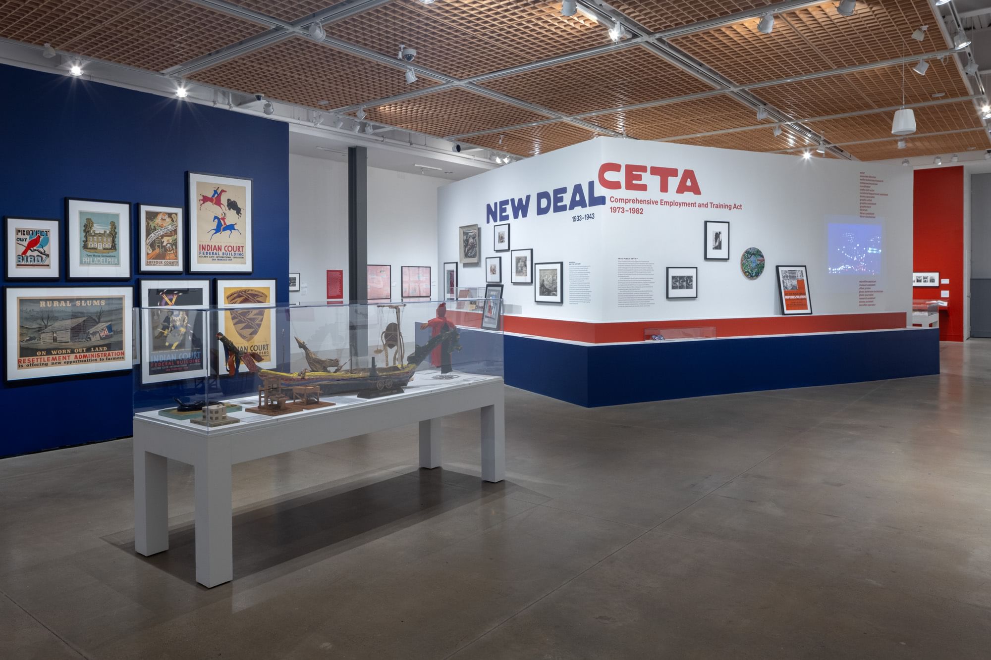 Large gallery with gray cement floor features many distinctive, colorful WPA-era posters on a dark blue wall at left.