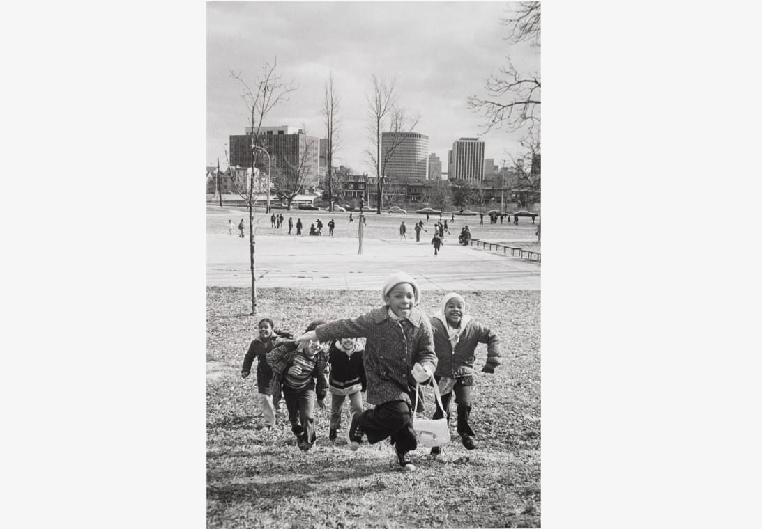 Vintage 1970s photo of five smiling children running up a small hill toward the viewer, a city skyline in the background.