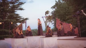 Atop a multi-tiered outdoor stage against a dimming evening sky, the actors pose in brown robes with yellow accents.