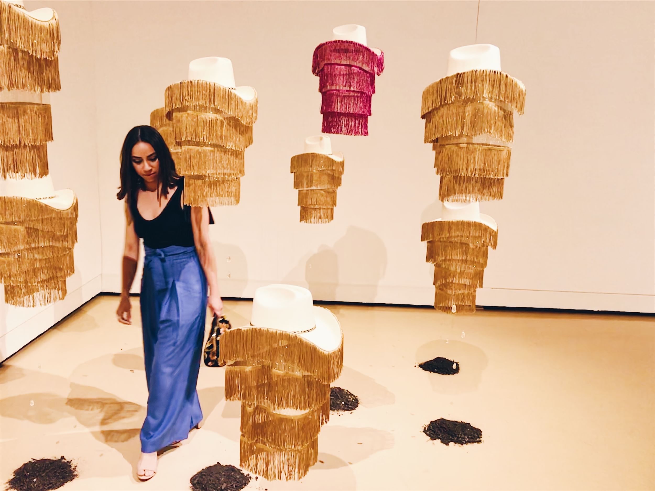 Vicky, wearing a black top & blue pants, walks under hats that drip with long fringe and hang from the ceiling of the gallery