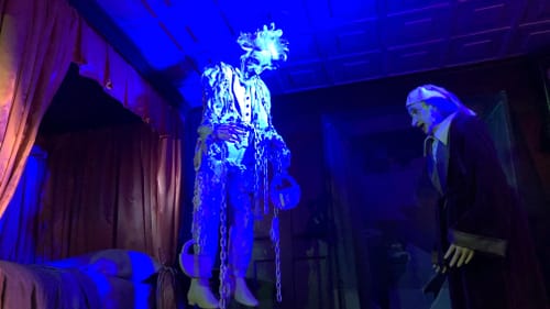 In a spooky blue-lit scene, Marley’s white ghost, covered in chains, floats above a terrified Scrooge in his nightgown.