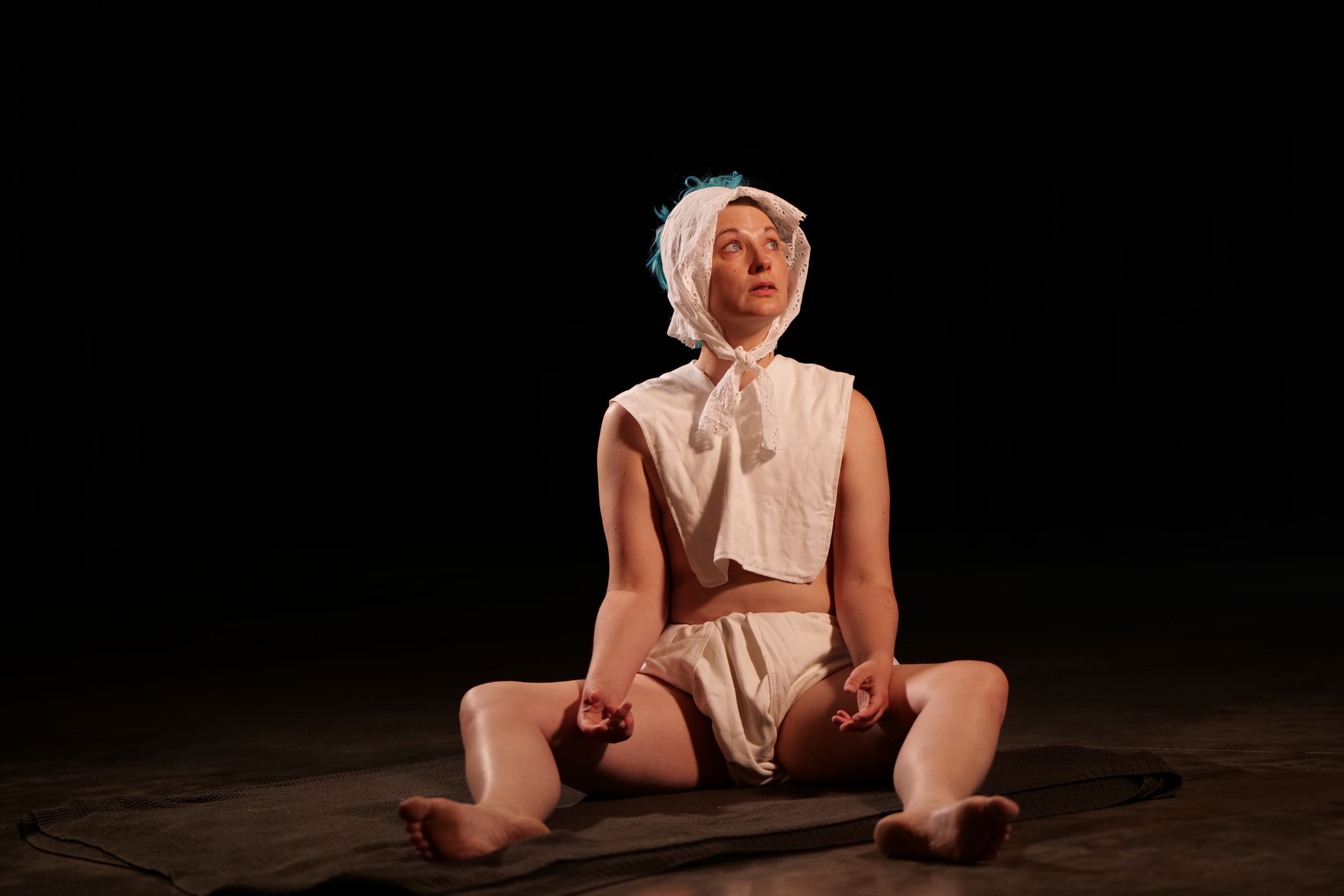 Tuomanen, a white person, sits on the floor like a baby, wearing a white kerchief, bib, and diaper.