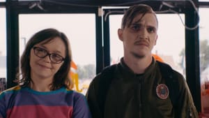 Two people look on, one smiles, the other looks unapproving or disgusted at something. They’re inside, a door behind them
