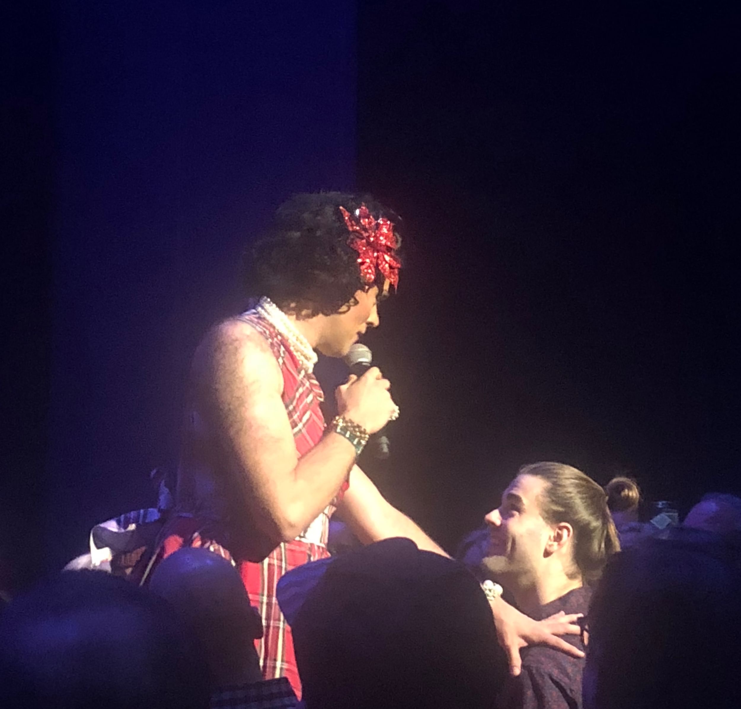 A tall, hairy drag queen in a red plaid dress touches the shoulder of a tall, smiling young man with a brown bun.