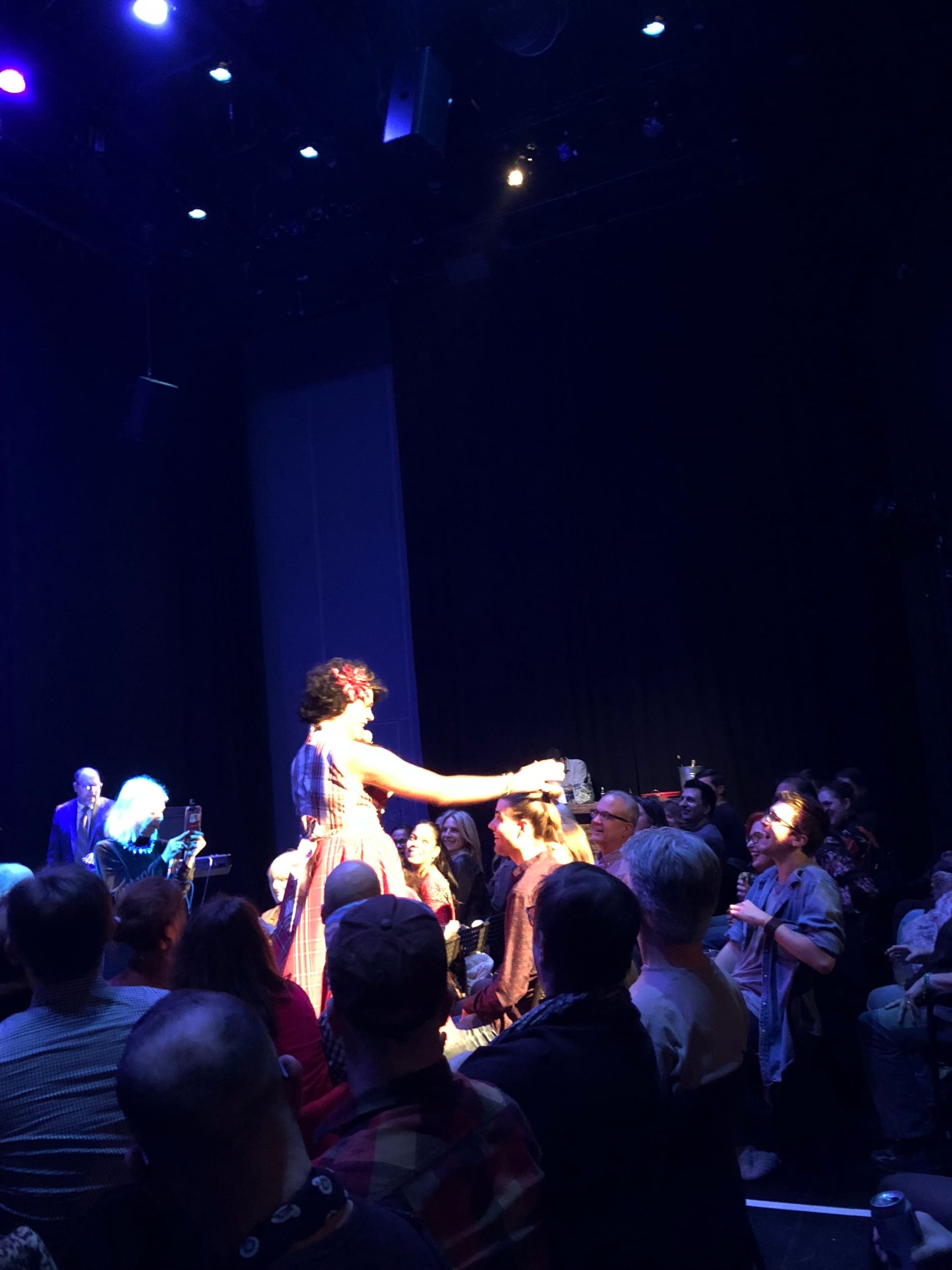 A tall, hairy drag queen reaches tenderly for a tall, smiling young man in the audience.