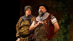 Lawton and Greer, with worried expressions, stand together in layered, detailed outfits reminiscent of vaudeville clowns