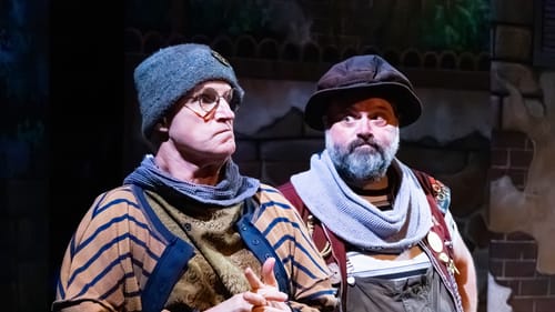 Lawton and Greer, with worried expressions, stand together in layered, detailed outfits reminiscent of vaudeville clowns