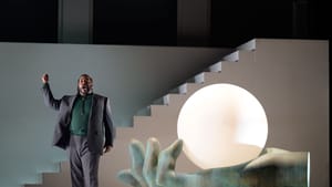 Outlaw, a Black man in a gray suit, sings with one fist in the air next to a giant hand sculpture holding a white orb