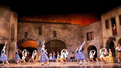 On a set that looks like a stone-walled courtyard with an orange sky, 25 dancers in period Spanish costumes pose joyfully