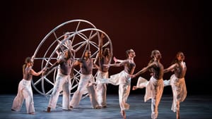 Three dancers in motion in front of a set of other dancers holding a large wheel-like structure, a dancer set inside