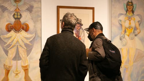 Seen from behind, two men look closely at some large Thrash works featuring fanciful, colorful full-body figures.
