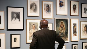 Seen from behind, a bald Black man in a suit jacket looks at a wall of many Dox Thrash portraits.