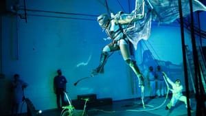 In dramatic blue lighting, actor Desirée Hall seems to fly in a dragon costume with giant wings, beside a cinderblock wall.