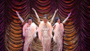 The Dreams pose with white-gloved arms up, against a luxe gold curtain, wearing silvery formal dresses and white feather boas