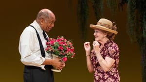 Hobbs, smiling and holding a flower bouquet, faces Scharfman, who smiles excitedly and wears a floral dress.