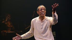 In loose pale cotton shirt and pants, X, a Black man, sits and gestures passionately mid-speech.