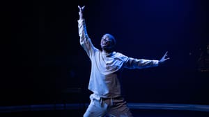 Washed in blue light, X kneels onstage with a desperate expression, arms extended upward beseechingly.
