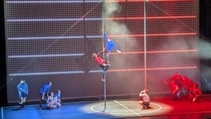 Two performers on a tall pole on stage in mid-movement. One in blue, the other red. Other performers in red and blue look on