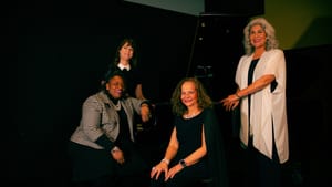 Four women in formal wear pose together in front of a piano in a dimly lit room.