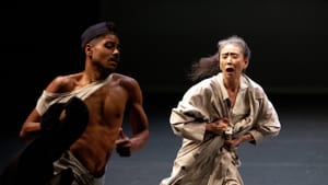 Jones and Otake in motion, looks like they're running in a dance movement, on a stage with a dark backdrop