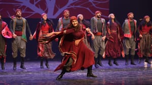 A line of dancers on stage, with one lead taking the front, dressed in thobes and head scarves