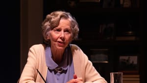 Reed, a white woman with short silver hair and a pale cardigan, speaks while sitting at a desk with her hands folded.