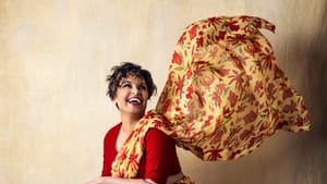 Shetty, an Indian woman, dressed in a yellow floral-print dress and red top, smiling looking up, in a portrait style photo