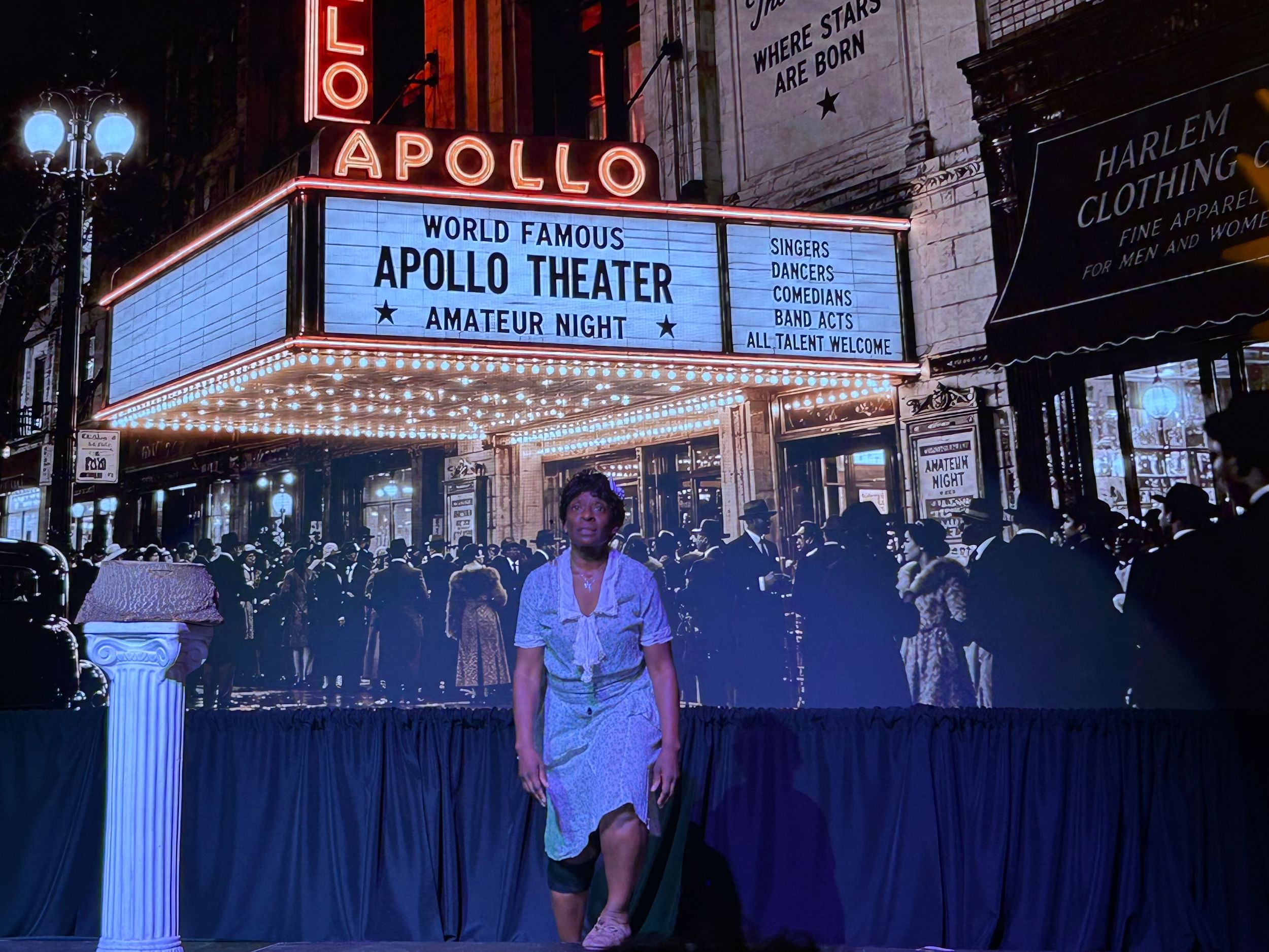 Scott, in a worn blue dress of early 1900s style, stands alone in front of a large projection of the Apollo Theater marquee.