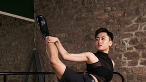 Godfrey, an Asian American dancer wearing black, strikes an angled pose with one leg extended, their hand on the ankle