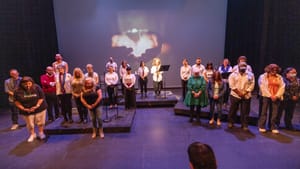23 storytellers of different ages, genders & races pose onstage with hands folded and heads lowered.