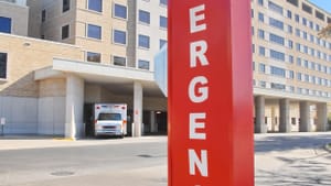 An exterior photo of a hospital with a large vertical sign that says “emergency” in red letters.