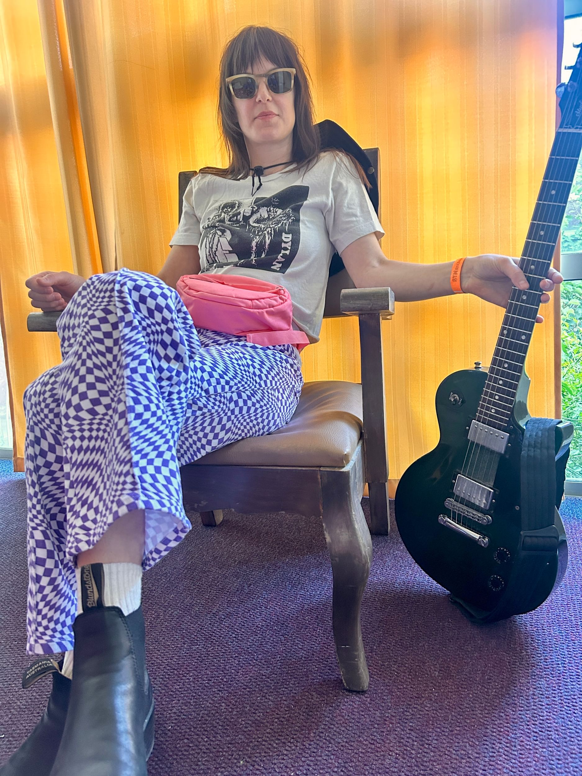 Low angle shot of a woman in a chair wearing sunglasses, a Bob Dylan shirt, checkered pants. A black guitar in her left hand