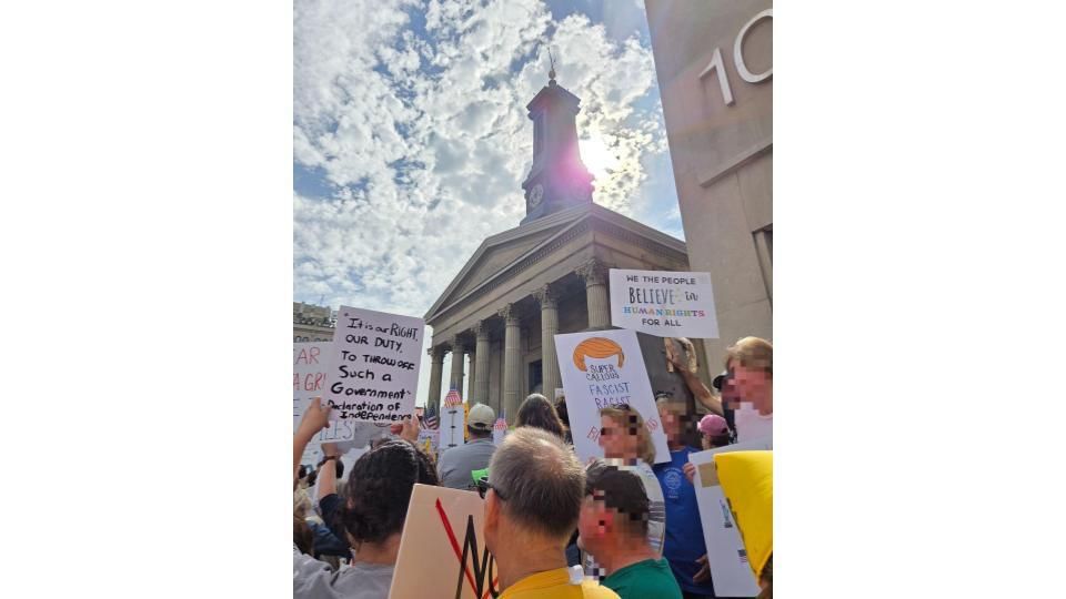 Protestors with handmade signs in a large crowd at the base of the pillared classical courthouse, sun shining through clouds