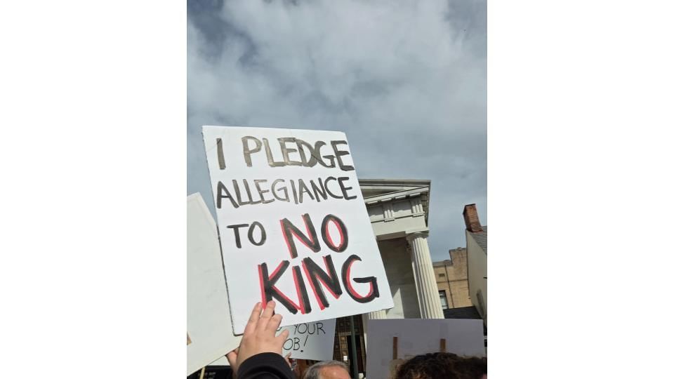 Close-up on a protestor outside the courthouse holding a hand-lettered sign that says “I pledge allegiance to NO KING"