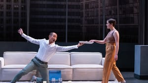 Makoto, an Asian man, kneels and flamboyantly holds out a plate to Claire, a white woman, in front of their large beige couch