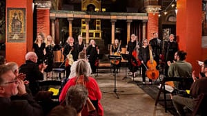 About 12 singers & musicians, wearing black, receive applause at the small, ornate venue with red brick pillars.