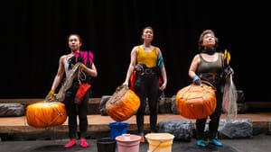 Three Asian women in free-diving gear stand determinedly in a row onstage, holding nets and large orange buoys.