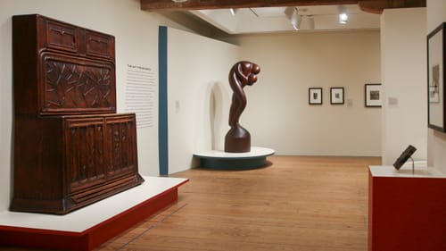 Gallery view of works including a huge, graceful wooden sculpture that resembles two people in a passionate embrace.