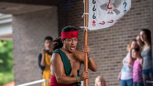 A person wears red and white clothes, bandana, and other clothing, carrying a flag (?) on a bamboo pole for a small crowd.
