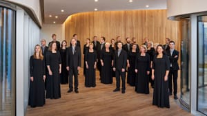 About two dozen performers in all black stand together in a hall room