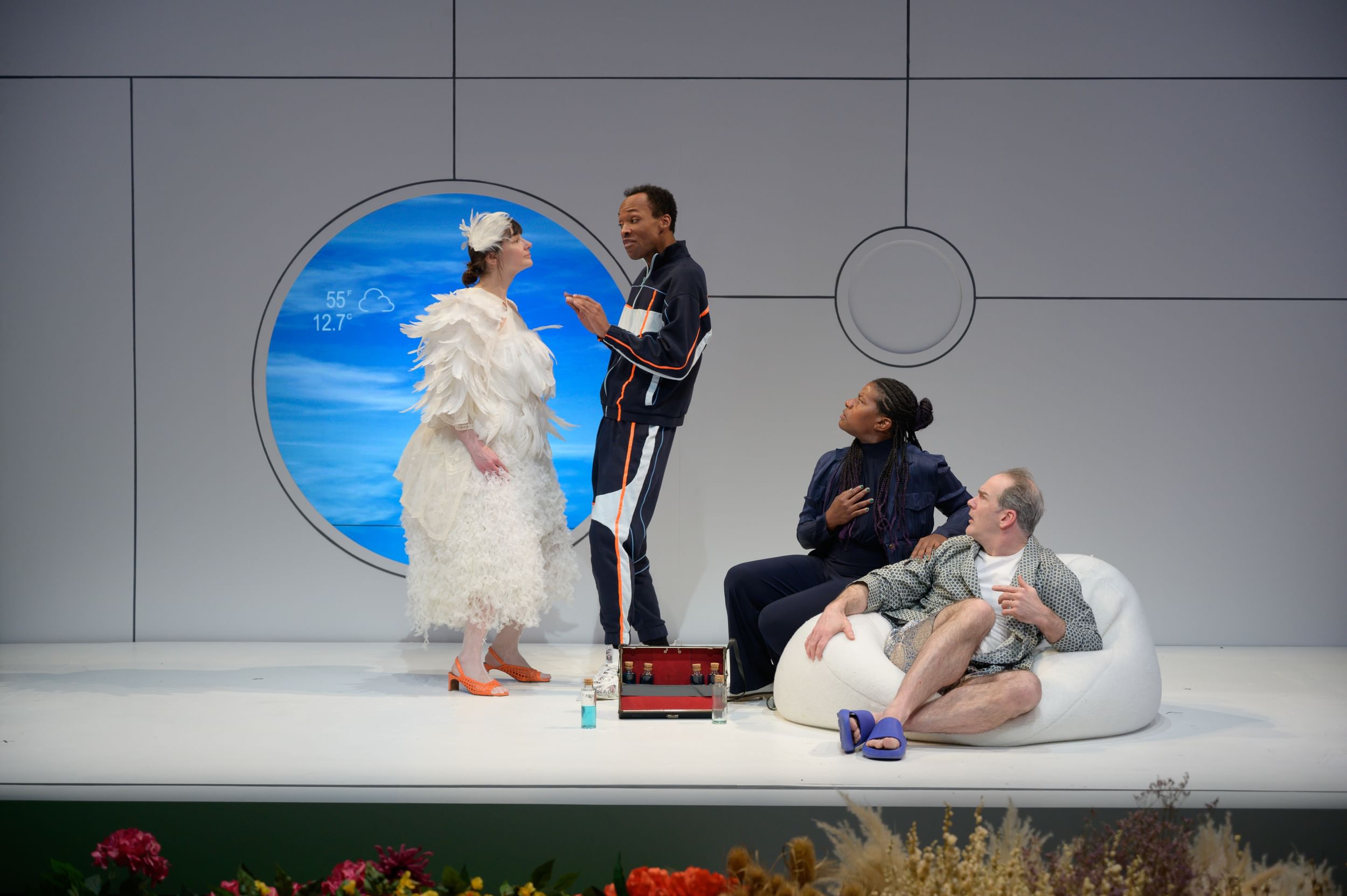 Gliko in white feathers, stands in front of a circular blue-lit window. Rishard & Kidwell sit on a beanbag looking at Pierce.