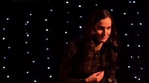 McCarthy onstage, leaning conspiratorially forward, with brown hair & sparkly top, against a black backdrop with white lights