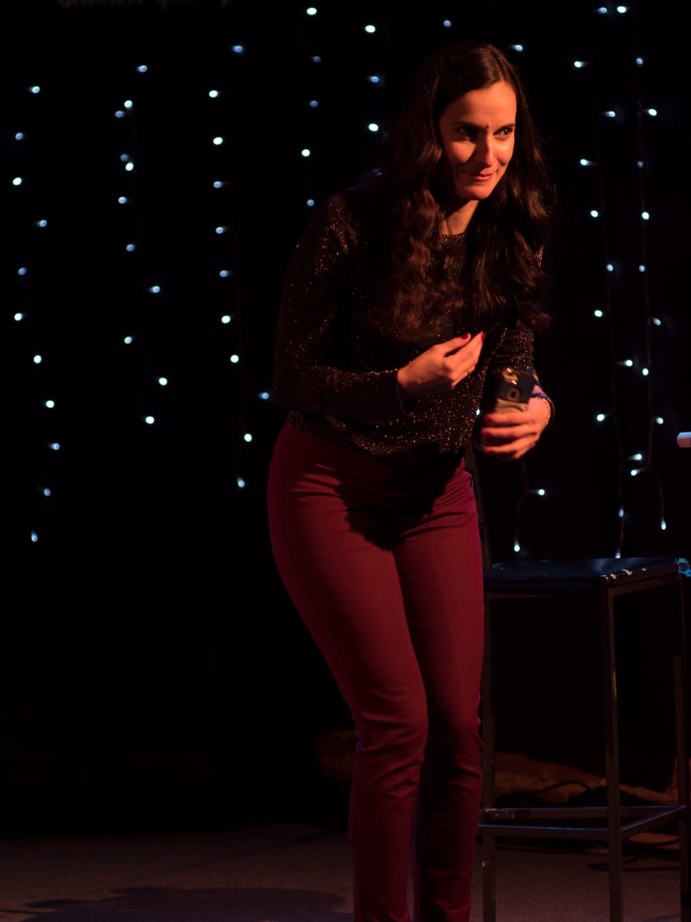 McCarthy onstage, leaning conspiratorially forward, with brown hair & sparkly top, against a black backdrop with white lights