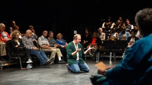 DelMarcelle, in a green cardigan and jeans, speaks while kneeling on the stage in front of the audience, seated around him.